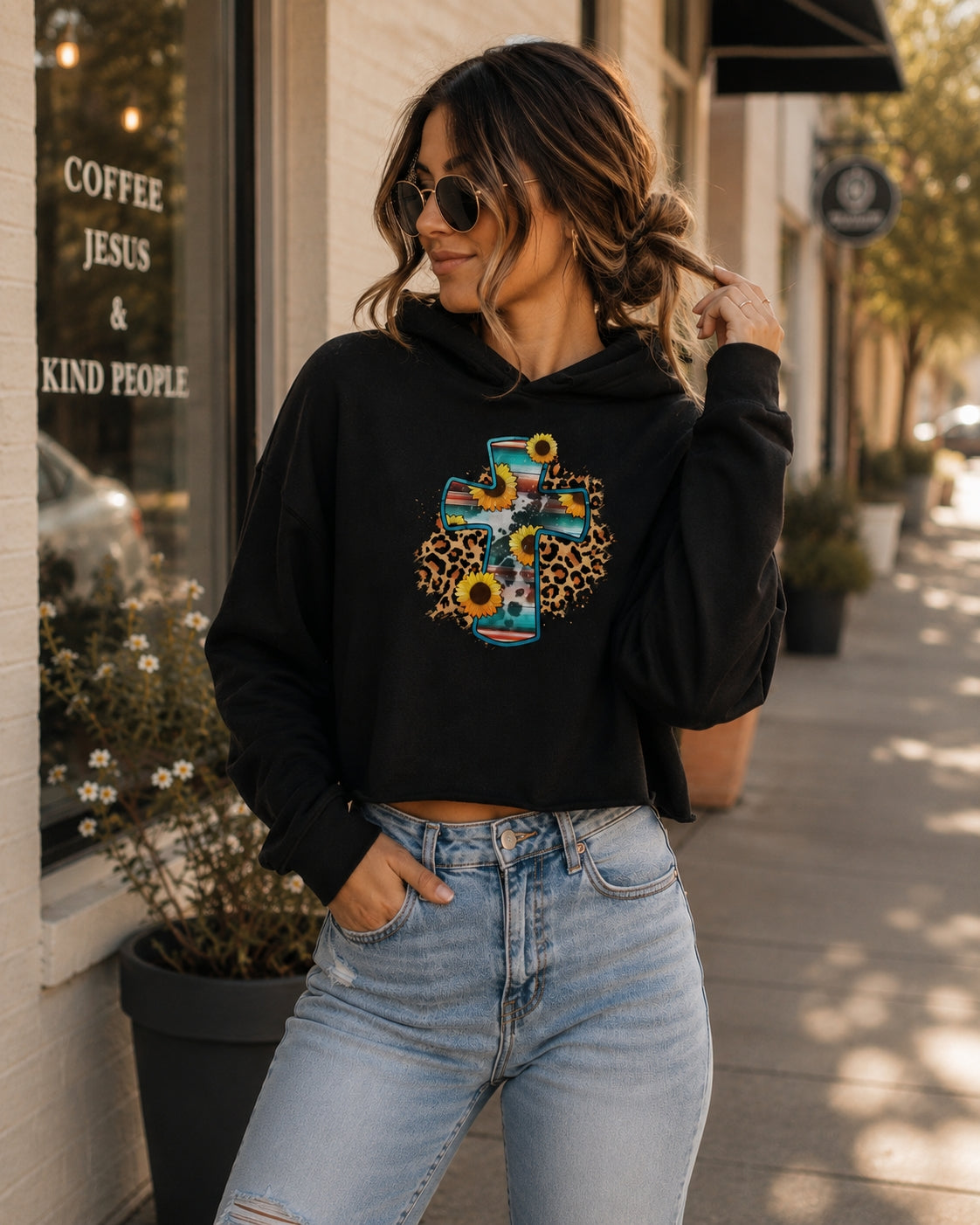 Woman wearing a black cropped Christian hoodie with a floral cross design while walking through a sunlit city street.