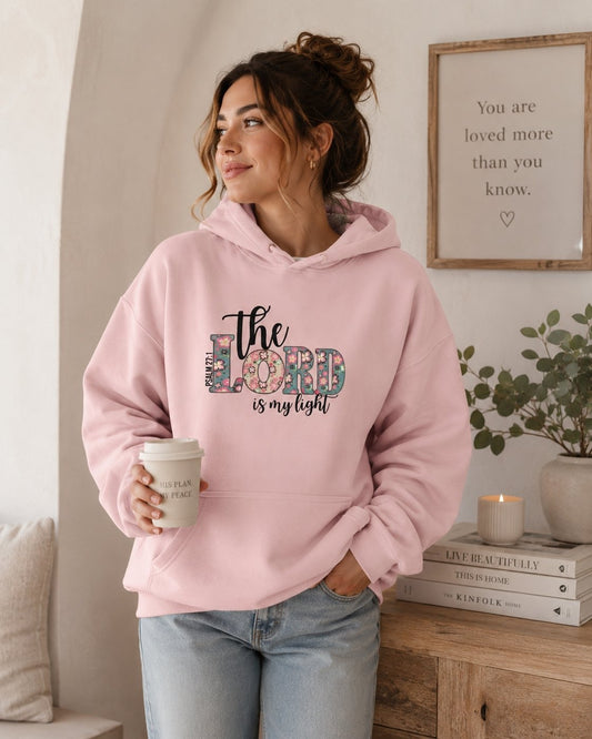Woman wearing The Lord Is My Light Psalm 27:1 light pink Christian hoodie in a cozy indoor setting.